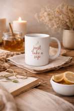 Load image into Gallery viewer, White ceramic wellness mug with “Just Take a Sip” text styled on a wooden surface with linen cloth, warm natural lighting, and minimal tea accessories in a calm self-care setting.